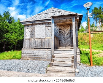 A rustic wooden cabin features a boarded window and wide steps, surrounded by green trees under a bright blue sky. - Powered by Shutterstock