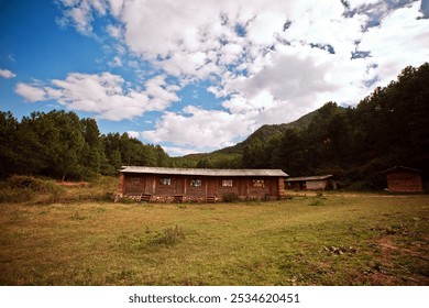 A rustic wooden cabin stands amid a lush green field, surrounded by dense trees and hills under a sky dotted with fluffy clouds. - Powered by Shutterstock