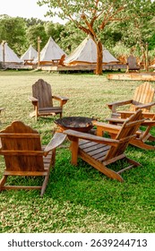Rustic wooden chairs around fire pit in scenic outdoor campsite with tents and lush greenery.