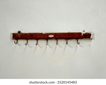 A rustic wooden coat rack mounted on a white wall. The coat rack has several metal hooks and shows signs of wear and tear, with chipped paint and a weathered appearance.  - Powered by Shutterstock