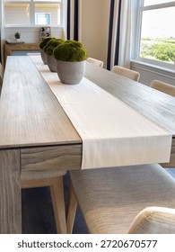 A rustic wooden dining table is styled with a white table runner and potted moss arrangements. The chairs are neutral-toned and the room is softly lit.