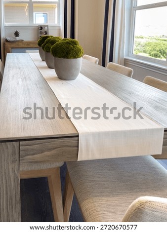 A rustic wooden dining table is styled with a white table runner and potted moss arrangements. The chairs are neutral-toned and the room is softly lit.