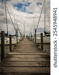 Rustic wooden dock stretches towards sailboats anchored at a tranquil marina. Partly cloudy sky adds a serene ambiance. Perfect for travel, leisure, and nautical themes.