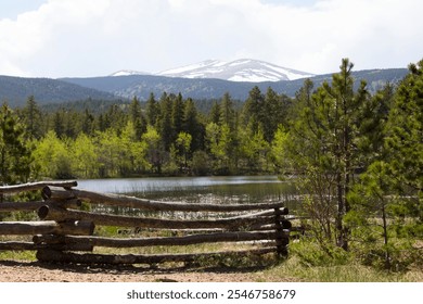 A rustic wooden fence frames a serene lake surrounded by lush greenery, with snow-capped mountains in the distance. - Powered by Shutterstock