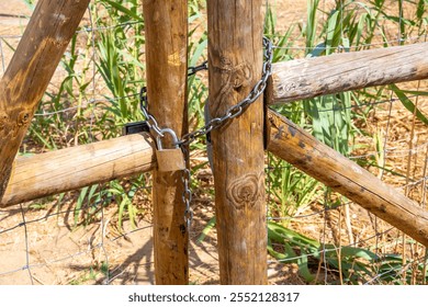 A rustic wooden gate with large wooden posts secured by a thick metal chain and padlock, with green plants visible through the wire mesh.