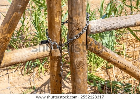A rustic wooden gate with large wooden posts secured by a thick metal chain and padlock, with green plants visible through the wire mesh.