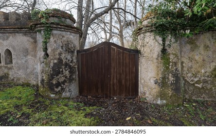 Rustic wooden gate set between aged stone pillars covered with moss and ivy, framed by bare trees in a quiet countryside setting with a timeless, slightly mysterious atmosphere.