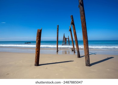 Rustic wooden pier remnants standing on a sandy beach with clear blue sky and calm ocean waves in the background. - Powered by Shutterstock