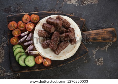 Rustic wooden serving board with grilled cevapi or cevapcici, tortillas and vegetables. Flatlay on a brown stone background