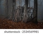 Rustic Wooden Shelter in Misty Autumn Forest. A rustic wooden shelter made of logs stands in a misty autumn forest. Surrounded by tall trees and fallen leaves, the scene evokes mystery and tranquility