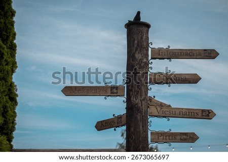 A rustic wooden signpost with multiple directional arrows in Latvian, pointing to local attractions or facilities such as “Informācija” (Information), “Noma” (Rental), “Viesu nams” (Guesthouse).