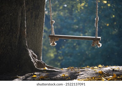 A rustic wooden swing hanging from a tree, surrounded by soft sunlight filtering through the leaves. The ground is covered with autumn leaves, creating a serene and peaceful atmosphere. - Powered by Shutterstock