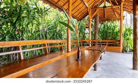 rustic wooden table with plant in bottle on it, surrounded by lush green plants and a thatched roof. - Powered by Shutterstock