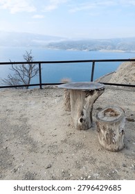 Rustic wooden table and stool sit on cliffside overlooking calm blue sea. Black metal railing protects viewpoint above sandy ground. Distant hills and coastal towns appear under clear sky