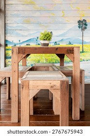Rustic wooden table and stools set with small green plant on balcony. Scenic mural wall background. Perfect for lifestyle, home decor, and cafe interior design themes.