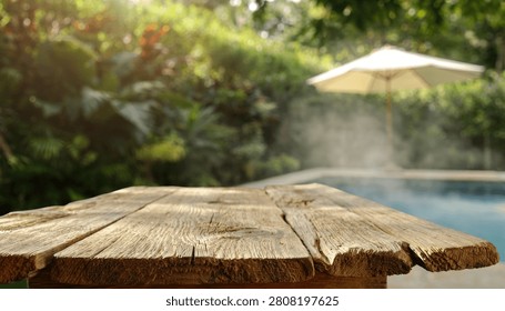 A rustic wooden table surface in the foreground with a blurred swimming pool and lush garden setting in the background on a sunny day.