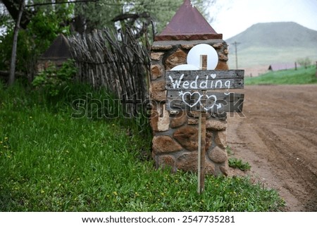 A rustic wooden wedding sign with heart decorations, directing guests down a dirt road toward a charming wedding venue, surrounded by natural greenery.