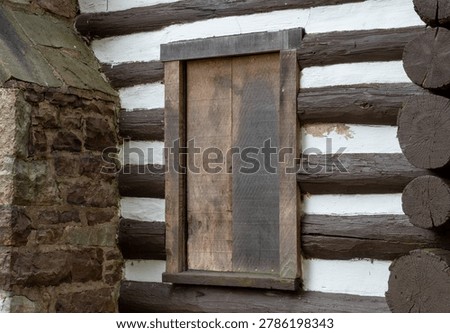 Rustic wooden window shutter on a historic log cabin wall with white chinking and natural stone foundation, showcasing traditional architecture and weathered textures.