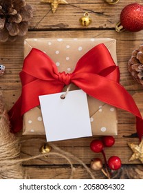 Rustic Wrapped Gift Box With Paper Gift Tag On A Brown Wooden Table With Red And Golden Christmas Decorations Around Top View. Winter Composition With Square Blank Gift Tag Mockup, Copy Space