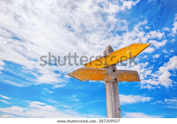 Rustic\
yellow crossroads sign against a cloudy blue\
sky.
