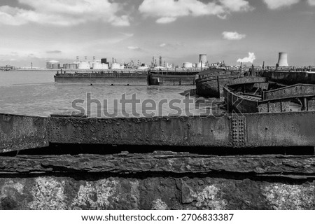 Rusting and decaying river barges along river estuary and modern oil refinery on the horizon, all under bright blue sky in Paull, Humberside, UK.