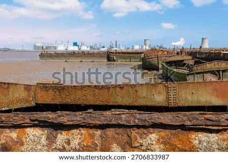 Rusting and decaying river barges along river estuary and modern oil refinery on the horizon, all under bright blue sky in Paull, Humberside, UK.