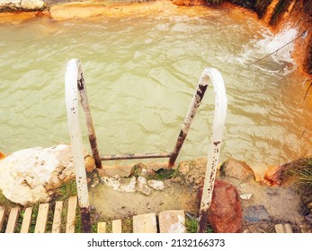 A Rusting Iron Staircase Leading To A Font With Muddy Mineral Water. Mountain Mineral Spring