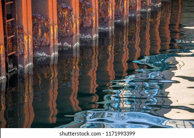 A Rusting Metal Sea Wall Provides A Nautical Backdrop For A Blue And Rust Reflection On Calm Water.