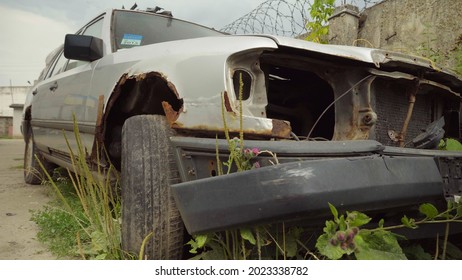 A rusty abandoned car in the parking lot, surrounded by a fence and barbed wire. A couple of cars are standing in a sump for automotive disassembly or metal processing. Restoration of a retro car.