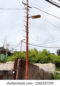 Rusty Abandoned Lamp Post. A Rusty Lamppost In A Green Village Against The Background Of A Concrete Foundation Of A Ruined House. Electrical Network Outside The City