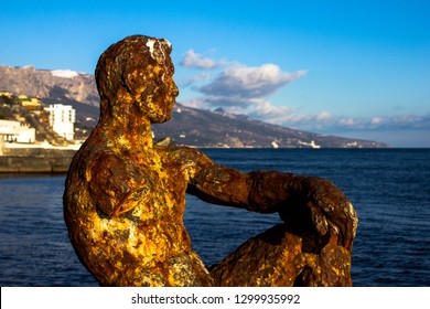 A rusty brown statue of a man falling apart and abandoned against the backdrop of nature, the sea and the mountains. Metal sculpture the whole covered with rust.