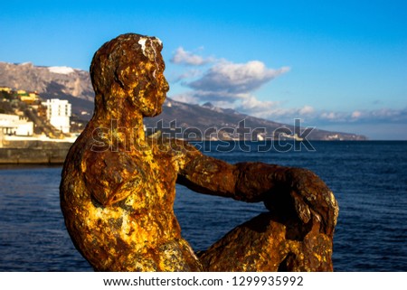 A rusty brown statue of a man falling apart and abandoned against the backdrop of nature, the sea and the mountains. Metal sculpture the whole covered with rust.