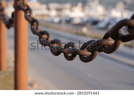 Rusty iron chain connecting two posts, creating a barrier alongside a road with blurred vehicles and buildings in the background, symbolizing restriction, strength, and old industrial elements