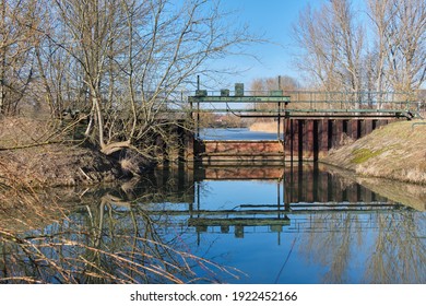 An Rusty Iron Little Weir Between Two Water Levels. The Upper Level Is Still Frozen, The Lower Lever Has Reflections