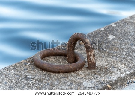 Rusty iron mooring ring fixed in a concrete quay beside calm blue water, creating a serene maritime still life. Light reflects softly off the rusted metal and rippling river