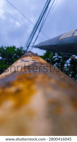 Rusty iron pillar with blurred background of green trees and power lines, showing urban decay and industrial elements.