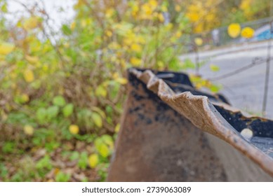 Rusty metal edge of an old object with textured surface in focus and blurred autumn foliage in the background creating a natural outdoor scene.