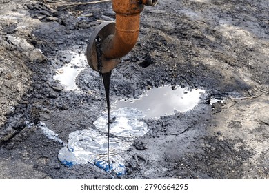 Rusty metal pipe leaking thick black oil onto the ground, creating a dark puddle. Concept of environmental pollution, toxic waste, and ecological disaster.