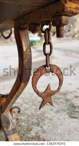 Rusty metal star shaped detail closeup. Turkish\
symbols star and moon hanging on rough rusty chain under rural wood\
wagon. Rusty metal texture. Old Turkish star and moon decorations.\
Vintage objects
