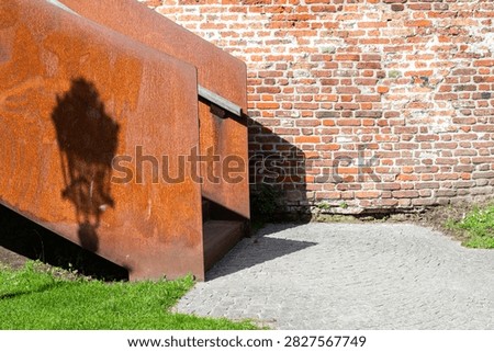 Rusty metal structure creates a shadow of a street lamp on a brick wall, with a cobblestone path and green grass in the foreground