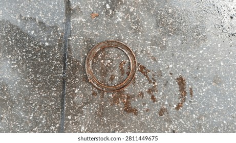 A rusty metallic ring or mechanical gasket piece lying on a wet, textured concrete ground with oil stains.