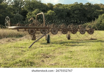 A rusty, old hay rake with multiple spiked wheels sits idle in a sunny, grassy field backed by dense, green trees.