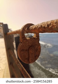 A rusty padlock hangs on a corroded railing overlooking the sea. The aged metal contrasts with the bright ocean view, evoking a sense of time passing and forgotten memories.

