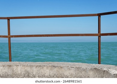 A rusty railing frames the tranquil turquoise water of a lake under a clear, blue sky. Gentle waves lap the shore.