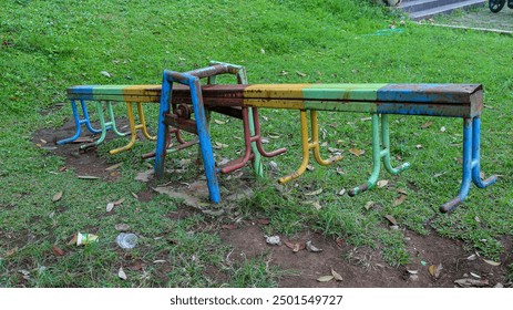 A rusty seesaw is abandoned in a grassy park. The metal structure is painted in various colors, faded with time. It stands as a reminder of past playtime and the passage of time. Rusty Seesaw. - Powered by Shutterstock