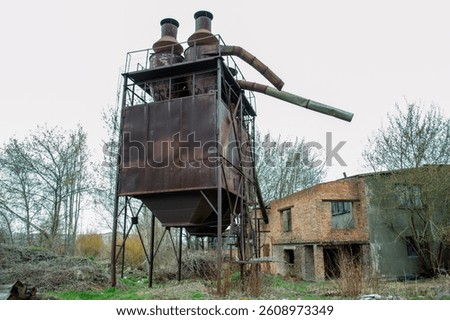 Rusty structure towers over wild grass and trees in an abandoned industrial site, showcasing a forgotten era of manufacturing and decay