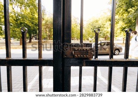 Rusty wrought iron gate with latch and vertical bars framing sunlit urban park, sidewalk, parked car, and distant pedestrian in warm afternoon light, with gate, latch, iron, metal detail