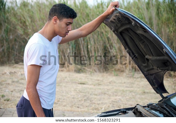 Rutinary car engine\
inspection check. Brown skin arabic young male standing on the road\
fixing car engine.