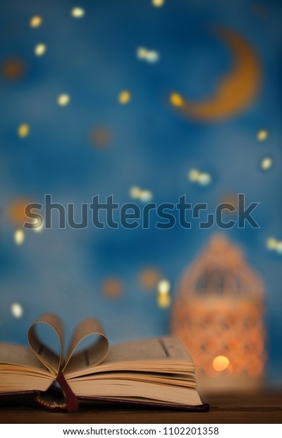 Sacred book of\
Quran on a wooden surface. Blue night sky with crescent moon and\
candle holder in the\
background