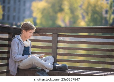 sad 11 year old child sitting on a bench in the park in autumn. a boy, a schoolboy, a teenager is waiting for someone on a bench outside, thinking, reflecting, reading something on a tablet. - Shutterstock ID 2365267847